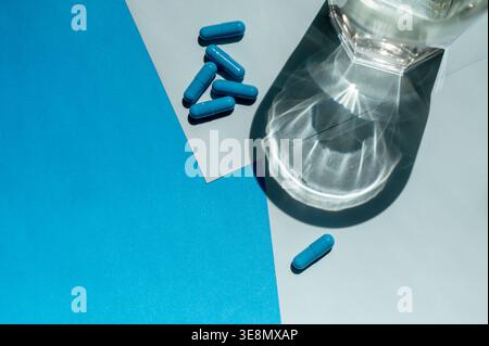 Eine einzige pharmazeutische Kapsel und mehrere Pillen auf einer blauen Studiooberfläche mit einem Glas Wasser. Konzept der Dosiskontrolle, Medikamentensicherheit und HE Stockfoto