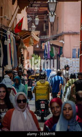 Lieferfahrer auf einem Motorrad, der durch eine geschäftige Marktstraße in der Medina von Marrakesch, Marokko, fährt Stockfoto