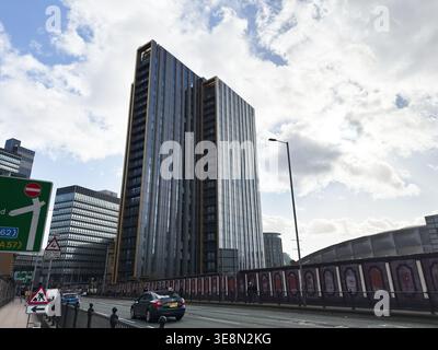 Manchester, Großbritannien - 22. Februar 2026: Hoher, moderner Turm der New Victoria Apartments mit Blick auf eine belebte Stadtstraße mit Autos, Schildern und dramatisch bewölktem Himmel Stockfoto