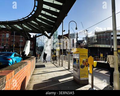 Manchester, Großbritannien - 22. Februar 2026: Pendler an einer stark frequentierten Straßenbahnhaltestelle in Manchester, Großbritannien, mit Fahrkartenautomaten, Shoppern und moderner Stadtarchitektur. Stockfoto