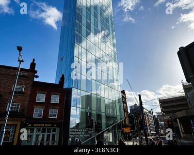Manchester, Großbritannien - 22. Februar 2026: Zeitgenössisches Glasgebäude, das Wolken über einer belebten Manchester UK Straße mit Fußgängern und Kränen reflektiert. Stockfoto