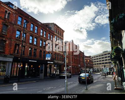 Manchester, Großbritannien - 22. Februar 2026: Historische Backsteinarchitektur säumt eine geschäftige Manchester UK Straße mit Geschäften, Autos und Fußgängern. Stockfoto
