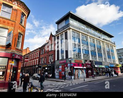 Manchester, Großbritannien - 22. Februar 2026: Fußgänger und Autos passieren lebhafte Ladenfronten unter einem modernen Bürogebäude in Manchester, Großbritannien. Stockfoto