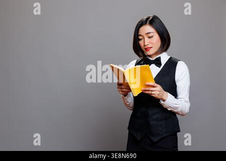 Nachdenkliche asiatische Kellnerin in professionellem Uniform-Lesebuch mit gelbem Einband. Junge attraktive Empfangsfrau, die Softcover-Tagebuch hält, während sie im Studio posiert Stockfoto