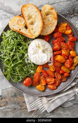Vorspeise mit Burrata, gebackenen Kirschtomaten und frischem Rucola in Nahaufnahme auf einem Teller auf dem Tisch. Vertikale Draufsicht von oben Stockfoto