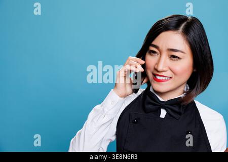 Hotel lächelnd fröhliche asiatische Frau an der Rezeption in Uniform, die auf dem Smartphone spricht. Fröhliche Kellnerin, die Anrufe entgegennimmt und mit dem Kunden des Catering-Service auf dem Mobiltelefon chattet Stockfoto
