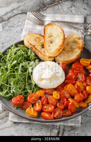 Vorspeise mit Burrata, gebackenen Kirschtomaten und frischem Rucola in Nahaufnahme auf einem Teller auf dem Tisch. Vertikal Stockfoto
