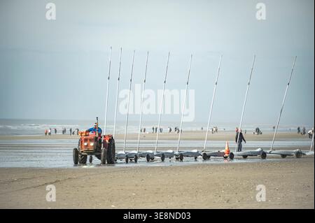 Quend-Plage, Frankreich - 4. April 2026: Ein roter Vintage-Traktor zieht eine lange Linie von Sandyachten über den breiten Sandstrand der Baie de Somme. Stockfoto