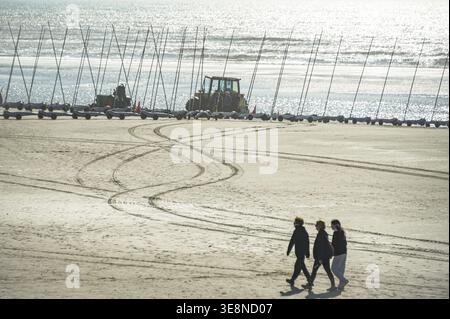 Quend-Plage, Frankreich - 4. April 2026: Die Menschen spazieren entlang der Küste an einer Reihe von Sandyachten und Traktoren vorbei, die am riesigen Strand unter strahlender Sonne geparkt sind. Stockfoto