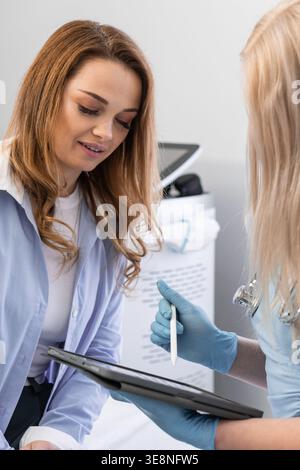 Weibliche Patientin hört aufmerksam einem Arzt zu, der medizinische Informationen mit einem Tablet in einer hellen Klinik erklärt. Der Gesundheitsdienstleister trägt Handschuhe und betont dabei Sicherheit und Professionalität Stockfoto