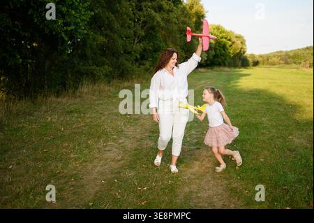 Glückliche Mutter und Tochter spielen mit Spielzeugflugzeugen auf dem Sommerfeld. Kaukasische Familie, die Spiele im Freien und Naturerholung genießt. Traum vom Flug, Kindheit und Bindungskonzept Stockfoto