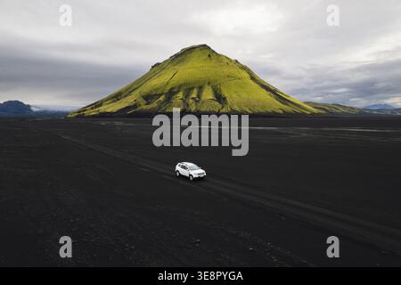 Aus der Vogelperspektive eines einsamen, weißen Fahrzeugs navigiert die krass schwarze Vulkanlandschaft in Richtung des leuchtend grünen moosigen Vulkans Mælifell Stockfoto