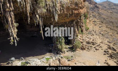 Aus der Vogelperspektive eines massiven Eingangs aus Kalksteinhöhle mit hängenden Stalaktiten in einer trockenen felsigen Landschaft; kleine Wanderer für Skala. Degub Cave, Sokotra Yeme Stockfoto