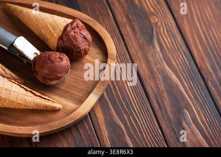 Rustikale Komposition mit Waffelkegeln und einer Schaufel mit Schokoladeneis im Löffel auf einem Holzteller. Stockfoto