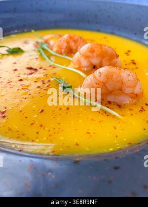 Eine leuchtend gelbe Suppe sitzt in einer blauen Schüssel. Drei Garnelen werden mit Pfeffer und kleiner grüner Garnitur darauf gelegt. Dieses Gericht wird duri genossen Stockfoto