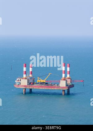 Luftaufnahme einer Offshore-Bauplattform mit rot-weiß gestreiften Türmen und einem gelben Kran auf dem blauen Meer in Taiwan. Stockfoto
