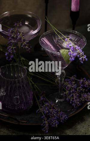 Zwei violette Lavendelcocktails in Martini-Gläsern und ein Glaskrug, garniert mit frischen Lavendelzweigen und Limettenscheiben, erscheinen auf einem dunklen rustikalen Stockfoto