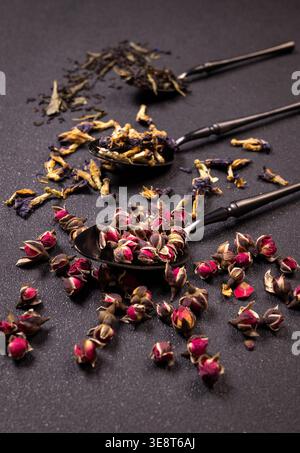 Verschiedene Zutaten für getrockneten Kräutertee, darunter Rosenknospen, Schmetterlingserbsenblüten und loser Blatttee, auf Vintage-Löffeln angeordnet und verteilt Stockfoto
