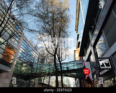 Manchester, Großbritannien – 28. Februar 2026: Eine Fußgängerbrücke aus Glas verbindet Bürogebäude und Geschäfte neben Bäumen und Reflexionen in Manchester, Großbritannien. Stockfoto