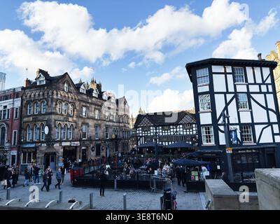 Manchester, Großbritannien - 28. Februar 2026: Geschäftiger Stadtplatz in Manchester, UK, mit Gebäuden im Tudor-Stil, Pubs, Sitzplätzen im Freien und Fußgängern. Stockfoto