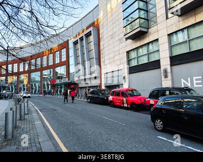 Manchester, Großbritannien - 28. Februar 2026: Fußgänger passieren geparkte Taxis vor einem geschwungenen Einkaufs- und Unterhaltungskomplex in Manchester, Großbritannien. Stockfoto