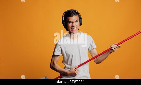 Asiatischer Sorgenloser, der Gitarre mit Besen spielt, sich vor der Kamera lustig macht und Musik hört. Fröhlicher, lustiger Mann, der lächelt und Lieder singt, ein Audio-Headset benutzt, Spaß mit Geräuschen zur Unterhaltung hat. Stockfoto