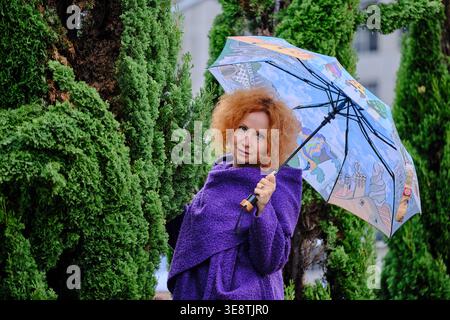 Wunderschöne rothaarige Frau mit durchsichtigem Regenschirm in einem üppig grünen Garten. Außenporträt bei Regen mit natürlichem Licht, weichem Fokus und vib Stockfoto
