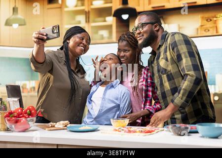 Die lächelnde Mutter nimmt Selfies mit der Familie auf dem Smartphone auf, während sie in der Küche köstliche hausgemachte Pizza kocht, während Kinder lachen und die Wärme der Bindung mit ihren Eltern genießen. Stockfoto