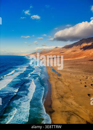 Blick aus der Vogelperspektive auf Playa del Confital mit türkisfarbenen Wellen, die auf dem Sandstrand und zerklüfteten vulkanischen Bergen unter einem blauen Himmel in Las Palmas krachen. Stockfoto