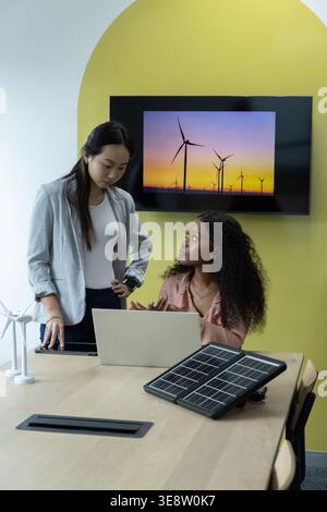 Verschiedene weibliche Kollegen in Blazer und rosa Bluse diskutieren über Solar-Panel-Wind-Modell-Laptop bei der Arbeit Stockfoto
