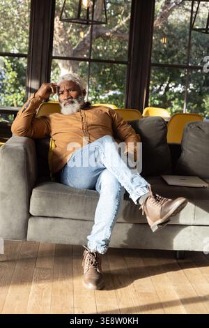Ranghoher Afroamerikaner sitzt auf grauem Sofa, trägt hellbraunes Hemd, Laptop in der Nähe Stockfoto