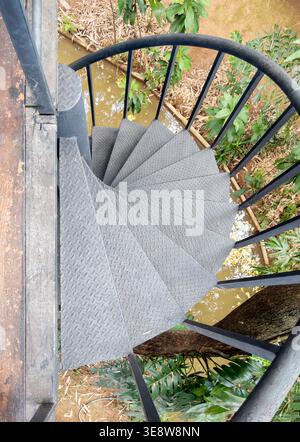 Hochwinkelaufnahme einer Wendeltreppe aus schwarzem Metall mit karierter Plattenstruktur. Industrielles Design über einem natürlichen Fluss in einem üppigen tropischen gar Stockfoto