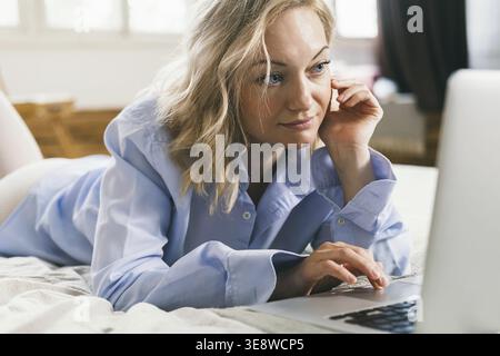 Ein Mädchen mit blonden Haaren liegt auf einem Bett vor einem Computer. Die Frau lächelt und kauft im Internet. Eine Frau arbeitet in ihrem Atelier. Cl Stockfoto
