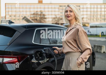 Laden eines Elektrofahrzeugs an einer öffentlichen Ladestation in einer städtischen Umgebung durch eine blonde Frau Stockfoto