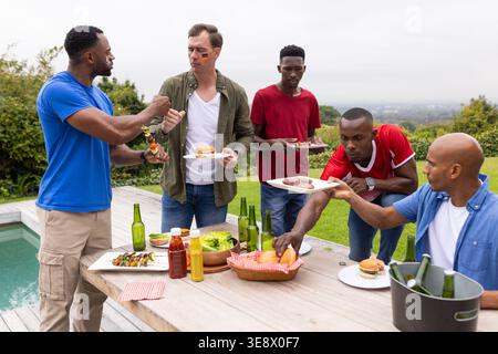 Verschiedene männliche Freunde grillen und teilen sich Burger auf Holzterrasse am Pool mit Bierflaschen Stockfoto