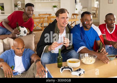 Verschiedene männliche Freunde schauen sich das Schlüsselspiel zu Hause an, indem sie sich beugen, Popcorn und Fußball erreichen Stockfoto