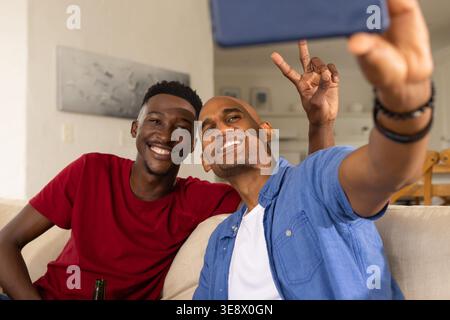 Afroamerikanische männliche Freunde, die rote und blaue Hemden tragen, sitzen auf dem Sofa und machen Selfie mit dem Telefon Stockfoto