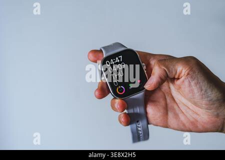 Eine Nahaufnahme einer Hand, die eine silberne Smartwatch mit einem grauen Silikonband vor einem neutralen hellen Hintergrund hält. Stockfoto