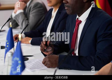 Nahaufnahme eines afroamerikanischen Geschäftsmannes, der eine Brille hält und an einem diplomatischen Treffen mit Kollegen am Konferenztisch teilnimmt. Geeignet für Regierungsrichtlinien Stockfoto