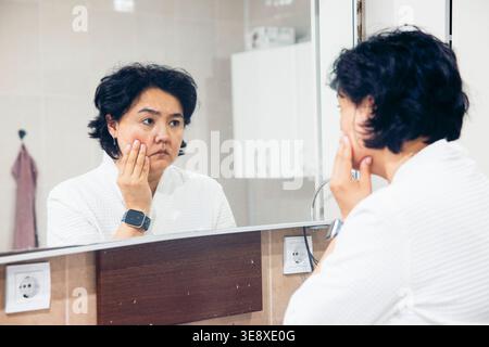 Traurige asiatische Frau mittleren Alters mit kurzen schwarzen Haaren untersucht ihre Haut in einem Badezimmerspiegel. Sie trägt ein weißes Gewand und berührt ihr Gesicht. Stockfoto