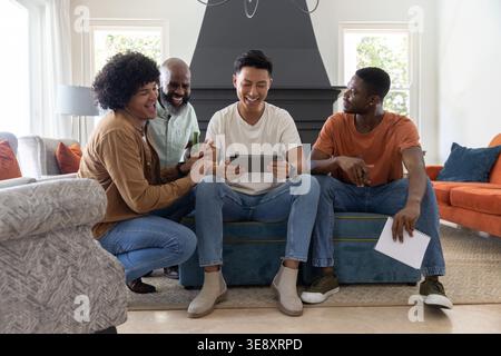 Verschiedene männliche Freunde, die sich im Wohnzimmer auf einen blauen Ottoman lehnen und ein Tablet mit Notebook-Flasche beobachten Stockfoto