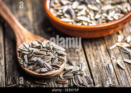 Nahaufnahme von gestreiften Sonnenblumenkernen, die in einem Holzlöffel mit einer Terrakotta-Schüssel auf einem verwitterten Holztisch gestapelt sind, was an eine rustikale Küche, einen gesunden Snack erinnert, Stockfoto