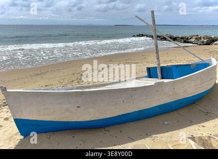 Ein hölzernes Fischerboot mit blauen Akzenten befindet sich am Sandstrand neben den Meereswellen und dem felsigen Ufer mit einem bewölkten Himmel über dem Meer Stockfoto