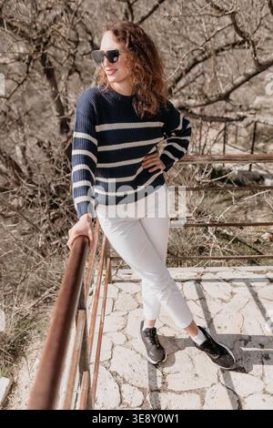Frau Outdoor Mode junge Frau in gestreiftem Pullover und Sonnenbrille lehnt sich im Frühjahr in einem Park auf ein Geländer Stockfoto