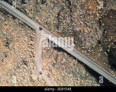 Drohnenblick aus der Luft mit Blick auf eine gerade Straße, die mit zwei Autos durch vulkanische Lavafelder führt, Lanzarote, Kanarische Inseln Stockfoto