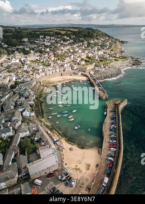 Aus der Vogelperspektive über den Hafen von Mousehole, Cornwall, mit türkisfarbenem Wasser, vertäuten Booten, Steinpiers und dem Dorf, das den Hügel hinaufklettert Stockfoto