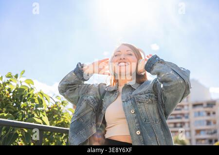 Glückliche junge Frau mit rotem Haar, die Musik über kabellose Kopfhörer hört, den urbanen Lebensstil genießt, die Augen geschlossen, an einem sonnigen Tag lächelnd Stockfoto