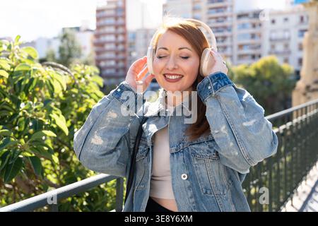 Frau trägt Kopfhörer, lächelt mit geschlossenen Augen, taucht draußen in Musik ein, während sie auf einer Brücke steht Stockfoto