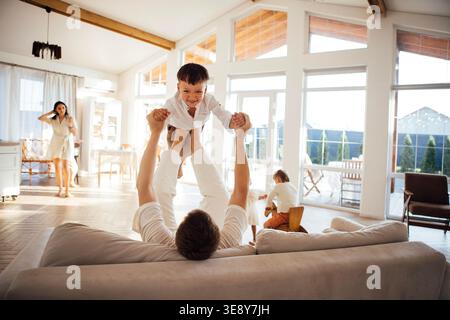 Vater hebt den Sohn spielerisch in ein helles Wohnzimmer, in dem die Familienmitglieder aktiv sind und eine fröhliche und liebevolle Familienumgebung präsentieren Stockfoto