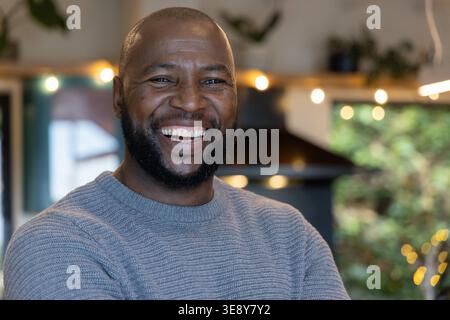 Afroamerikaner lächelt mit gekreuzten Armen in der Küche, trägt einen grauen Pullover und Lichterketten. Bärtig, Porträt, Modell, Strickwaren, gemütlich, warm, ungekühlt Stockfoto